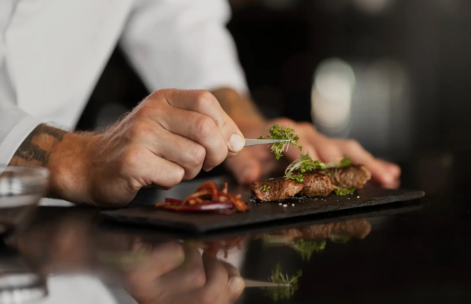 Chef profissional a empratar carne grelhada em pedra ardósia, adicionando microervas frescas com pinça – gastronomia premium e alta cozinha.
